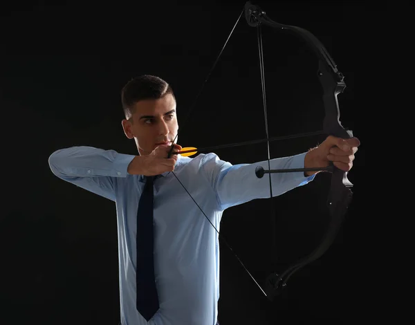 Joven hombre de negocios practicando tiro con arco sobre fondo oscuro — Foto de Stock