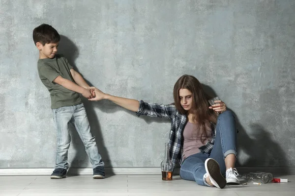 Женщина пьет алкоголь, в то время как маленький сын тянет руку к серой стене — стоковое фото