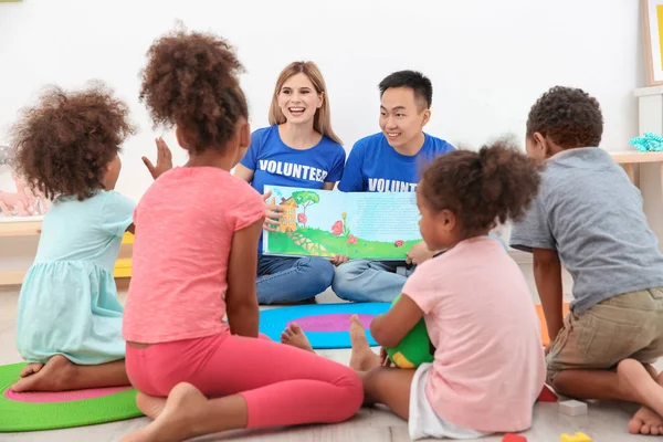 Jonge vrijwilligers lezen boek met kleine kinderen in lichte kamer. Vrijwilligerswerk in het buitenland concept — Stockfoto