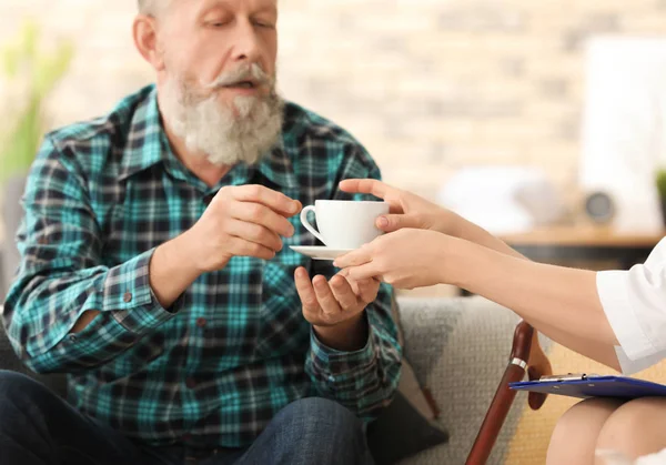 Cuidador joven dando taza de té a un hombre mayor en casa — Foto de Stock