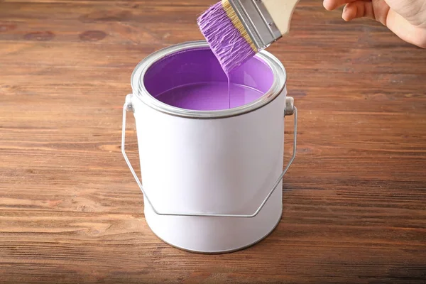 Woman dipping brush into tin can with violet paint on wooden background