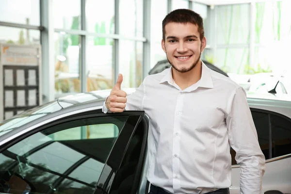 Handsome car salesman showing thumb up gesture near automobile in dealership centre — Stock Photo, Image