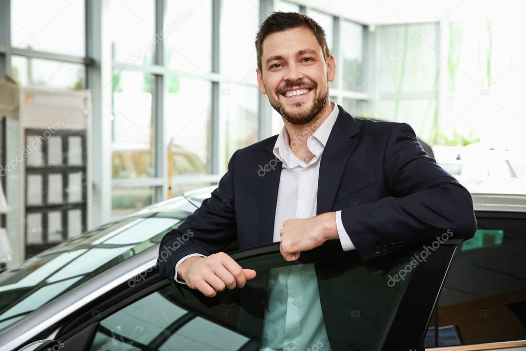 Handsome car salesman standing near automobile in dealership centre