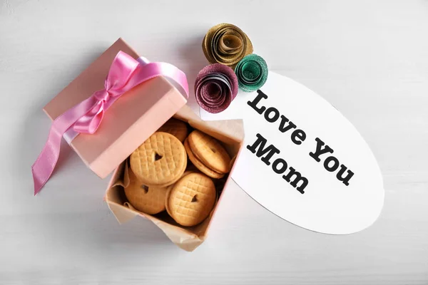 Note with words "Love you mom" and box of delicious cookies on light background — Stock Photo, Image