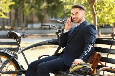 Bisiklet sürdükten sonra parkta bankta oturan genç yakışıklı işadamı cep telefonuyla konuşuyor.