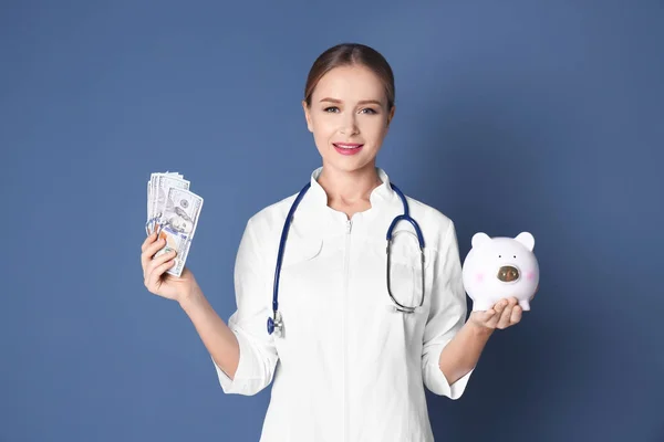Mladá lékařka drží peníze a prasátko na barvu pozadí — Stock fotografie