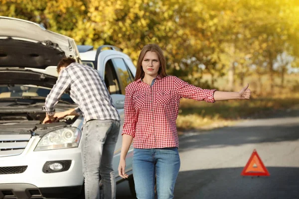 Młoda kobieta autostopem na drodze, podczas gdy człowiek stara się naprawić uszkodzony samochód — Zdjęcie stockowe
