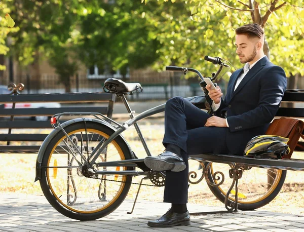 Joven hombre de negocios guapo con teléfono móvil y bicicleta sentado en el banco en el parque — Foto de Stock