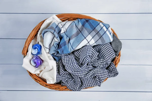 Basket with dirty clothes and laundry pods on white wooden background, top view — Stock Photo, Image