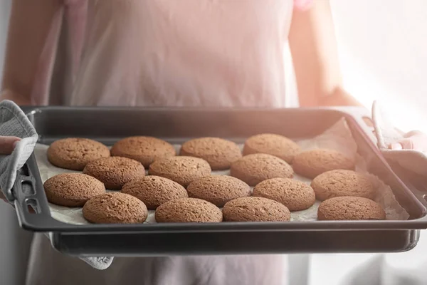 Woman holding baking tray with delicious oatmeal cookies in kitchen, closeup