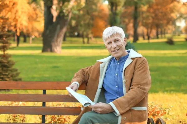 Стильный зрелый мужчина, сидящий на скамейке и читающий газету в осеннем парке — стоковое фото