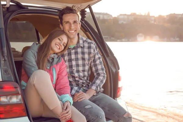 Hermosa pareja joven sentada en el maletero del coche cerca del río —  Fotos de Stock