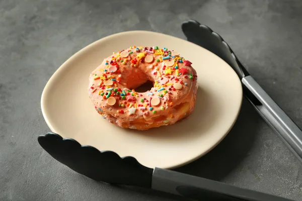 Plate with yummy colorful donut and tongs on grey background — Stock Photo, Image