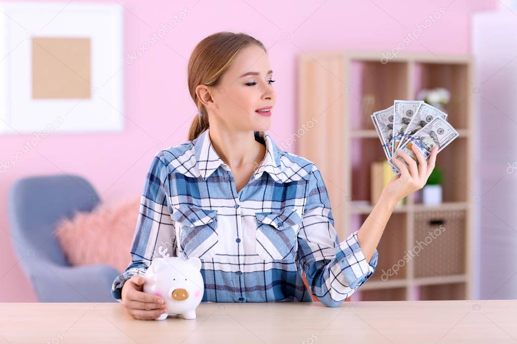 Young woman with piggy bank and money at table indoors