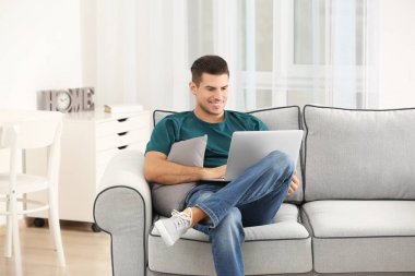 Evde koltukta istirahat ederken dizüstü bilgisayar kullanan yakışıklı adam