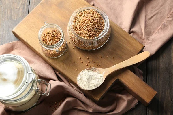 Harina de trigo sarraceno y granos crudos en utensilios de cocina sobre tabla de madera — Foto de Stock