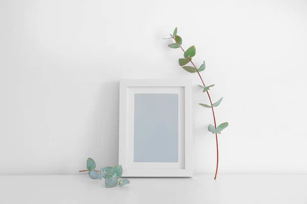 Empty frame and eucalyptus branch on table near white wall