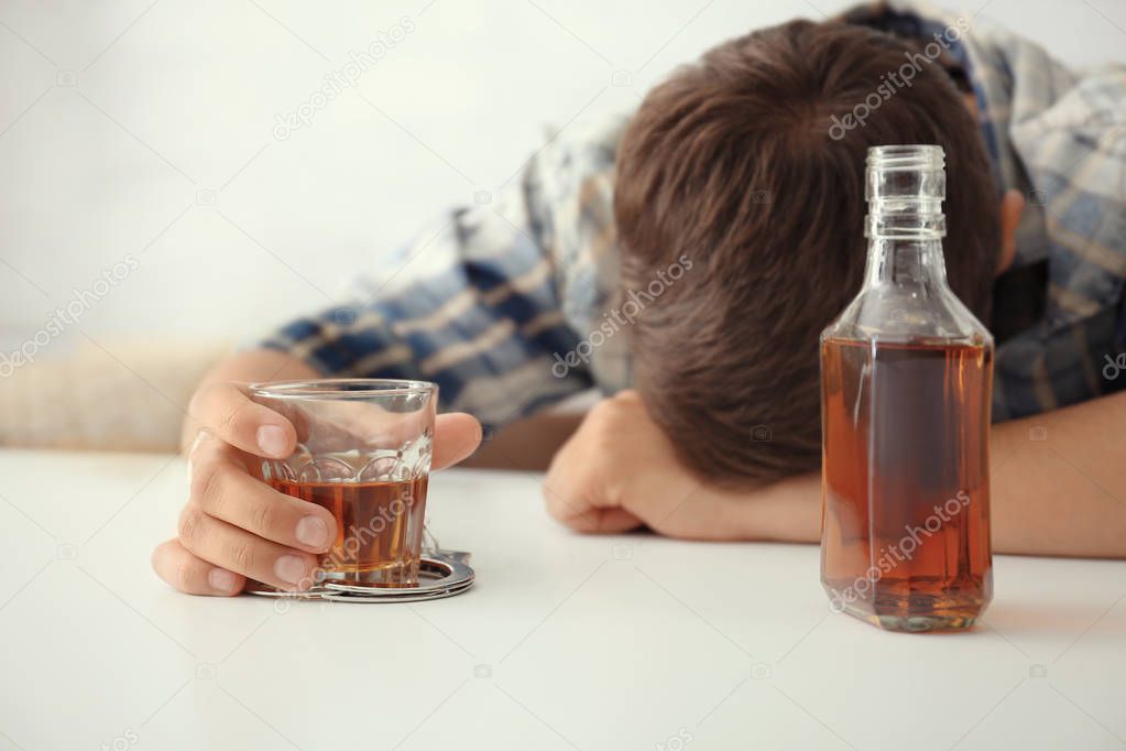 Drunk man in handcuffs with glass and bottle of alcohol sitting at table indoors. Alcohol dependence concept