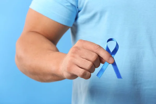 Young man holding blue ribbon on color background, closeup. Prostate cancer awareness concept