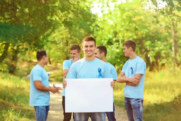 Νεαρός άνδρας σε t-shirt με μπλε κορδέλα κρατώντας πανό κενό σε εξωτερικούς χώρους. Έννοια συνειδητοποίησης καρκίνου του προστάτη — Φωτογραφία Αρχείου