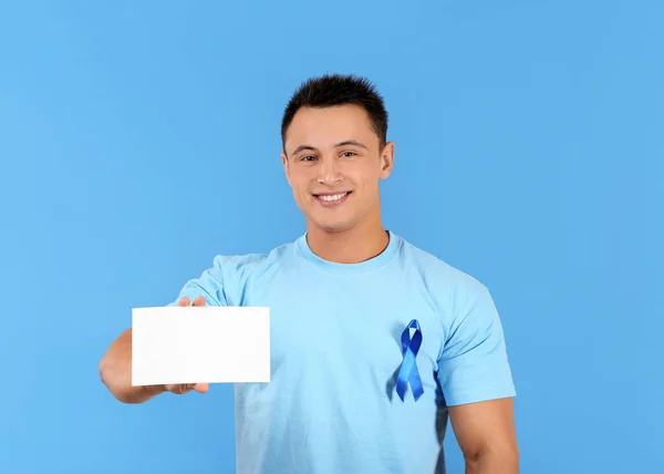 Hombre joven en camiseta con cinta azul sosteniendo la tarjeta en blanco en el fondo de color. Concepto de conciencia del cáncer de próstata —  Fotos de Stock