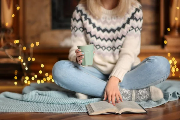 冬の休日の飾られた居間で本を読みながらリラックスできるお茶のカップを持つ若い女性 — ストック写真
