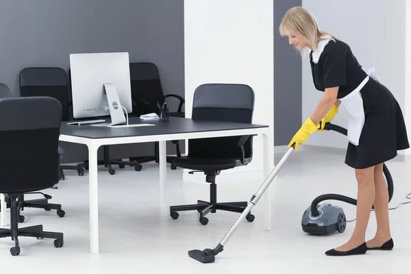 Mature charwoman hoovering floor in office with vacuum cleaner — Stock Photo, Image