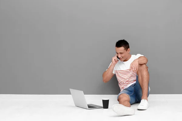 Jeune homme avec ordinateur portable et téléphone portable assis sur le sol près du mur gris — Photo