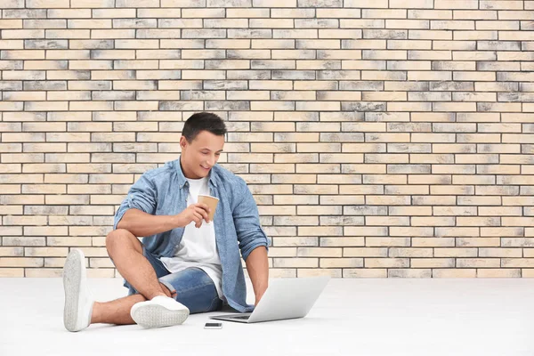 Młody człowiek z laptopa i kawę siedząc na podłodze w pobliżu mur z cegły — Zdjęcie stockowe