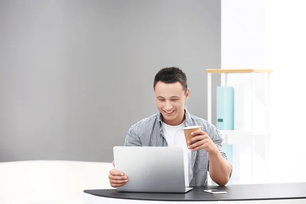 Νεαρός άνδρας, πίνοντας καφέ και να χρησιμοποιούν φορητό υπολογιστή στο γραφείο — Φωτογραφία Αρχείου