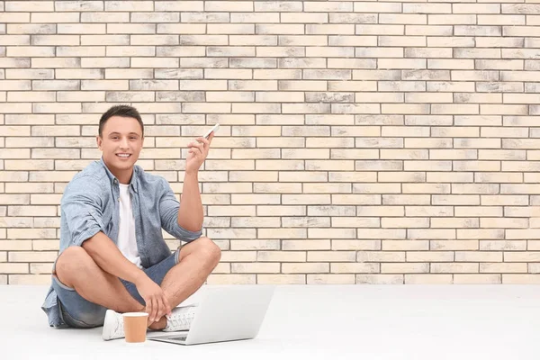 Młody człowiek z laptopa i telefon komórkowy siedząc na podłodze w pobliżu mur z cegły — Zdjęcie stockowe