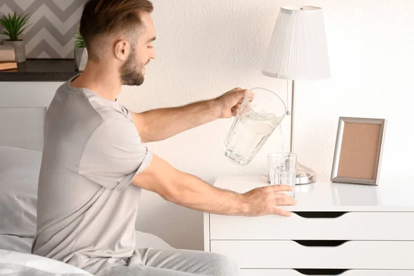 Mañana de joven guapo vertiendo agua en el vaso en casa — Foto de Stock