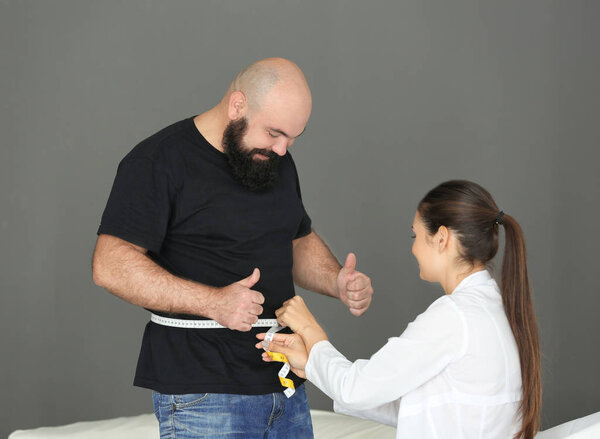 Young female doctor measuring waist of overweight man with centimeter in clinic
