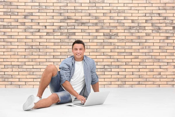 Happy young man with laptop sitting on floor near brick wall