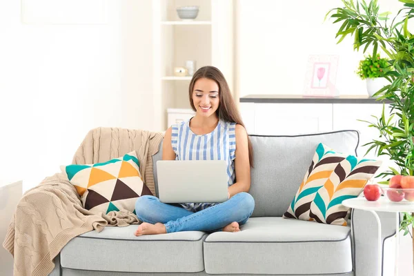 Jovem mulher usando laptop enquanto se senta no sofá acolhedor dentro de casa — Fotografia de Stock