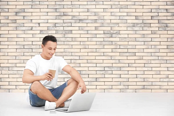 Młody człowiek z laptopa i kawę siedząc na podłodze w pobliżu mur z cegły — Zdjęcie stockowe