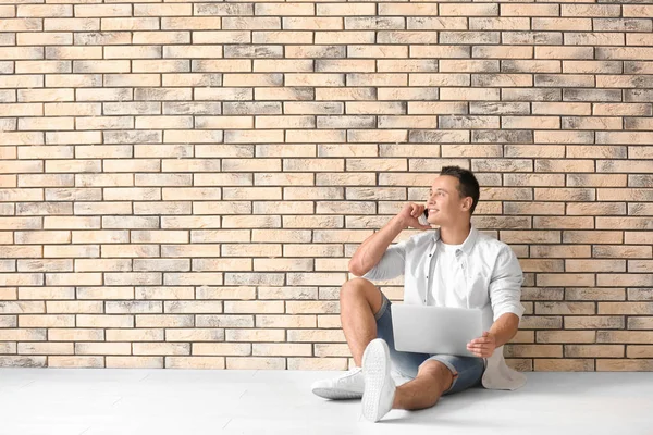 Молодий чоловік з ноутбуком і мобільним телефоном сидить на підлозі біля цегляної стіни — стокове фото