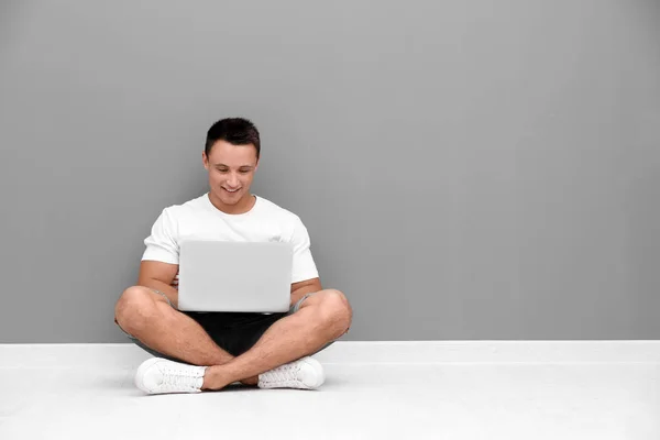 Hombre joven con portátil sentado en el suelo cerca de la pared gris —  Fotos de Stock