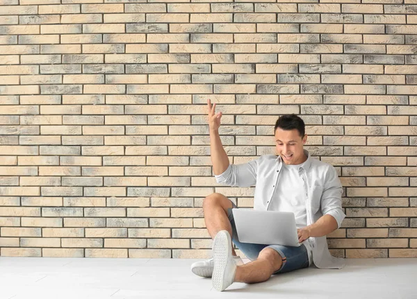 Szczęśliwy młody człowiek z laptopa siedząc na podłodze w pobliżu mur z cegły — Zdjęcie stockowe