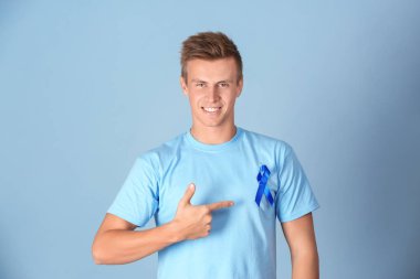 Young man pointing at blue ribbon on his t-shirt against color background. Prostate cancer awareness concept clipart