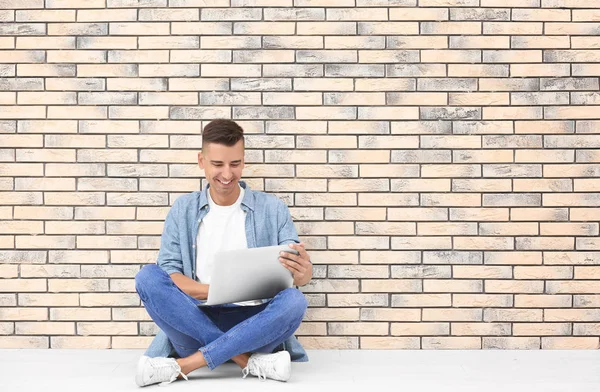 Happy young man with laptop sitting on floor near brick wall