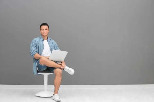 Młody człowiek z laptopa siedząc na krześle koło szare ściany — Zdjęcie stockowe
