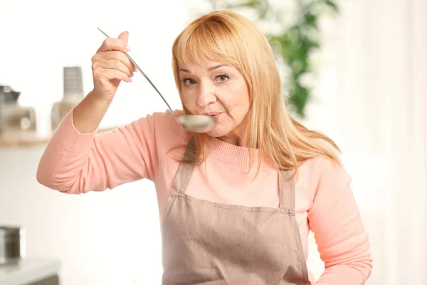 Beautiful mature woman tasting dish while cooking in kitchen — Stock Photo, Image