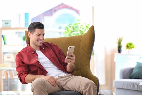 Красивый молодой человек с телефоном сидит в удобном кресле дома — стоковое фото