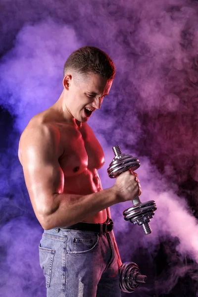 Muscular young bodybuilder with dumbbells in colorful smoke