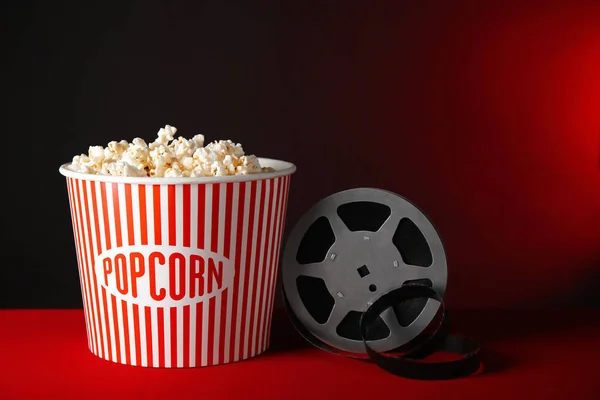 Striped bucket with tasty popcorn and movie reel on table against dark background