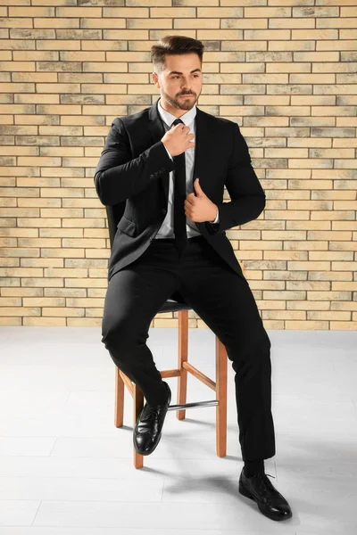 Bonito homem em terno formal sentado na cadeira contra a parede de tijolo — Fotografia de Stock