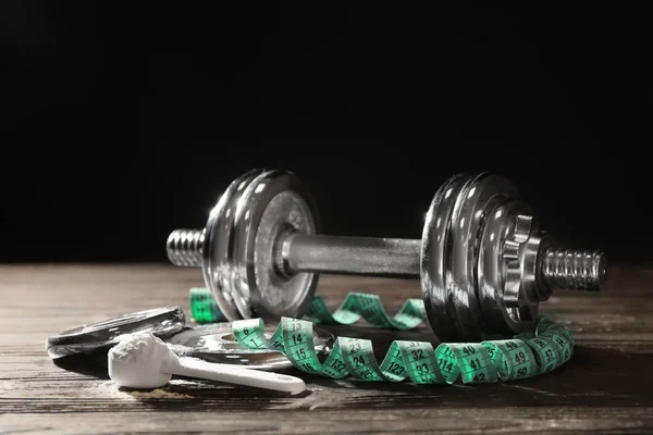 Composition with protein powder, measuring tape and dumbbell on table against dark background