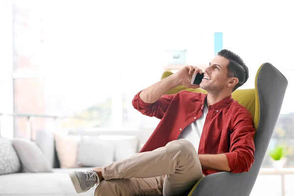 Pohledný mladý muž, zatímco sedí v pohodlné křeslo doma mluví o telefonu — Stock fotografie