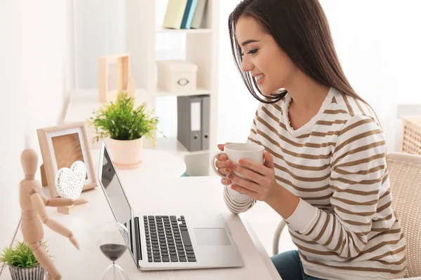 Junge Frau trinkt Kaffee, während sie mit modernem Laptop am Tisch arbeitet — Stockfoto